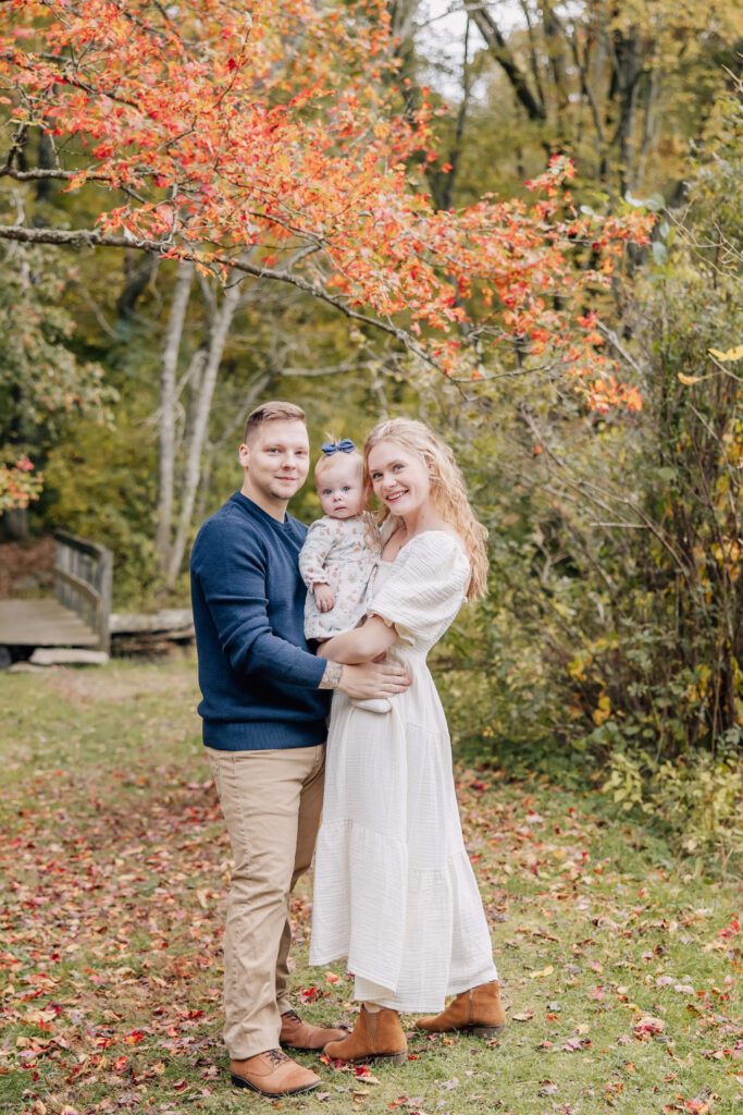Mother, baby, and father pose together in front of a wooden bridge and a bright orange tree during peak fall foliate at Ledyard Sawmill.